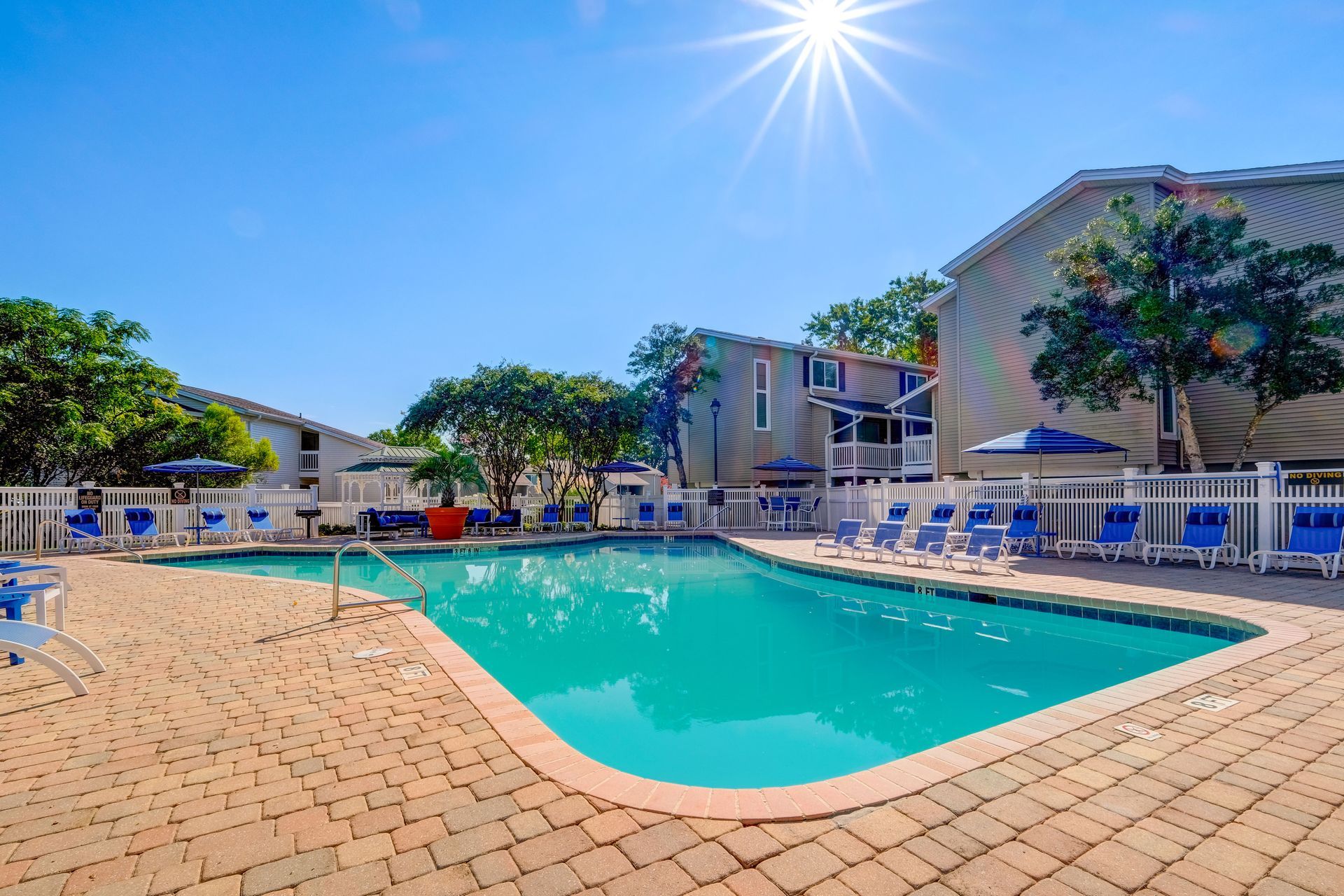 A turquoise swimming pool surrounded by a brick patio, lounge chairs, and apartment buildings under a sunny blue sky.