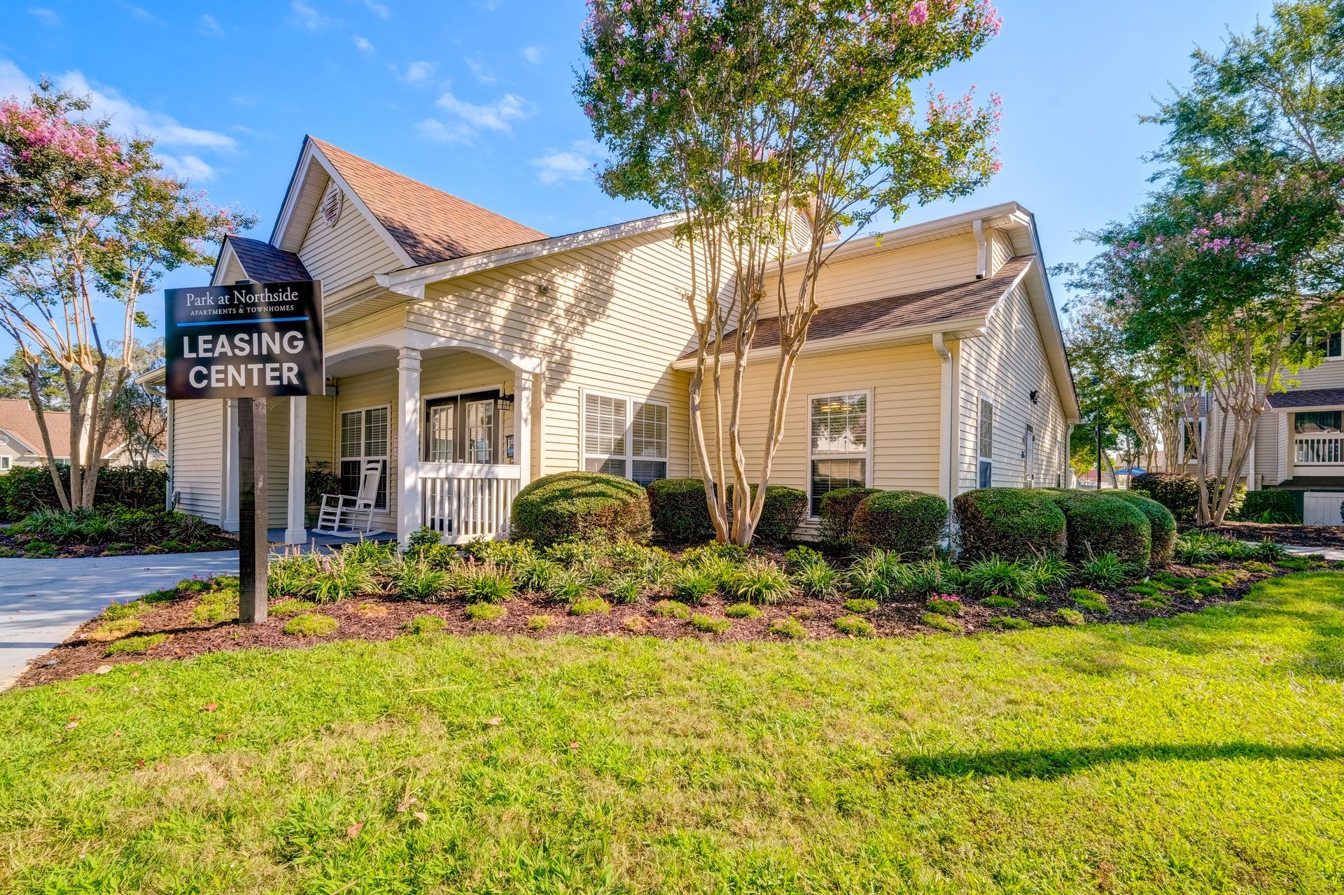 A beige leasing center building surrounded by manicured shrubs, green grass, and trees under a clear blue sky.