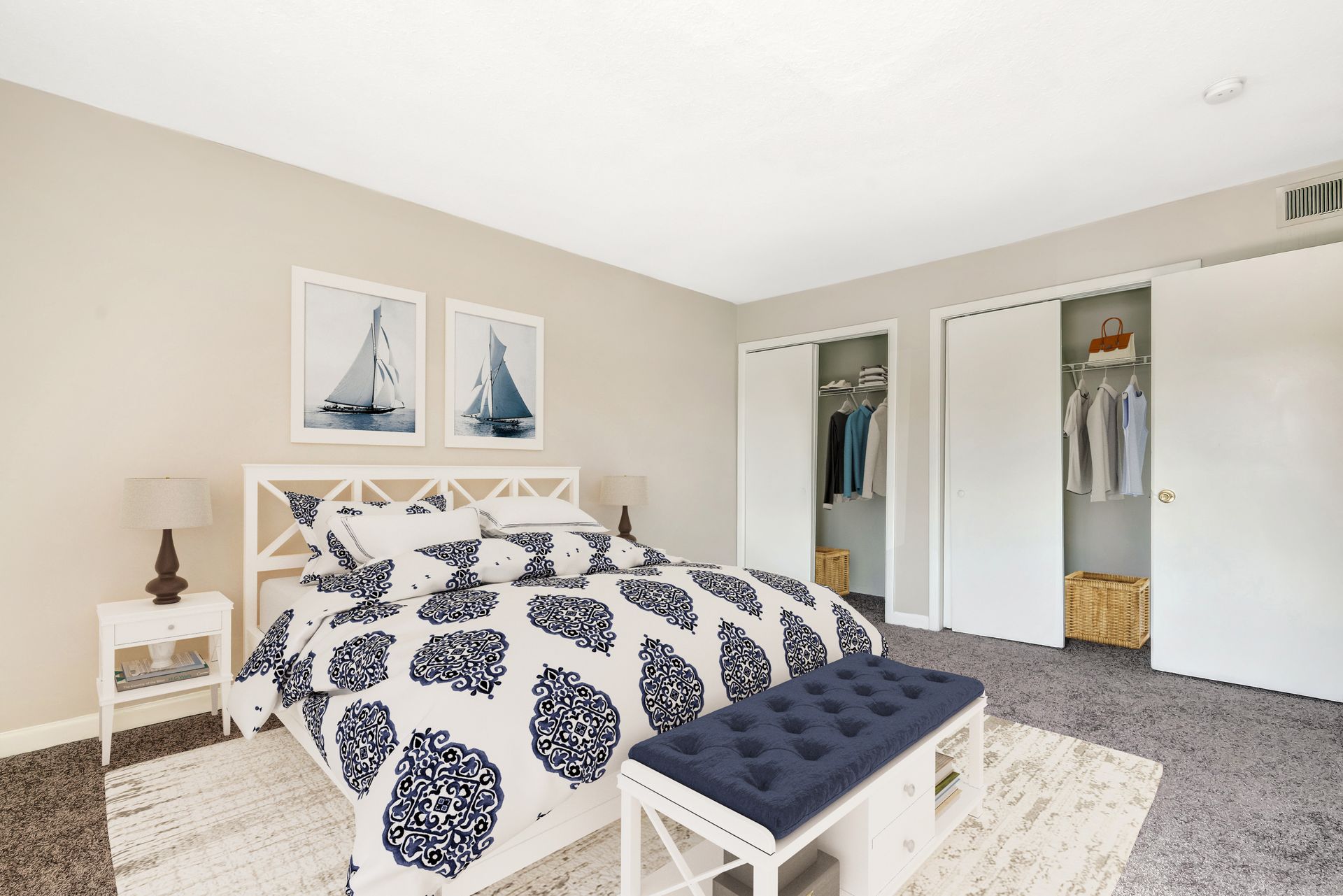 A bright bedroom features a queen bed with a blue and white patterned duvet, a matching bench, and two white closets.