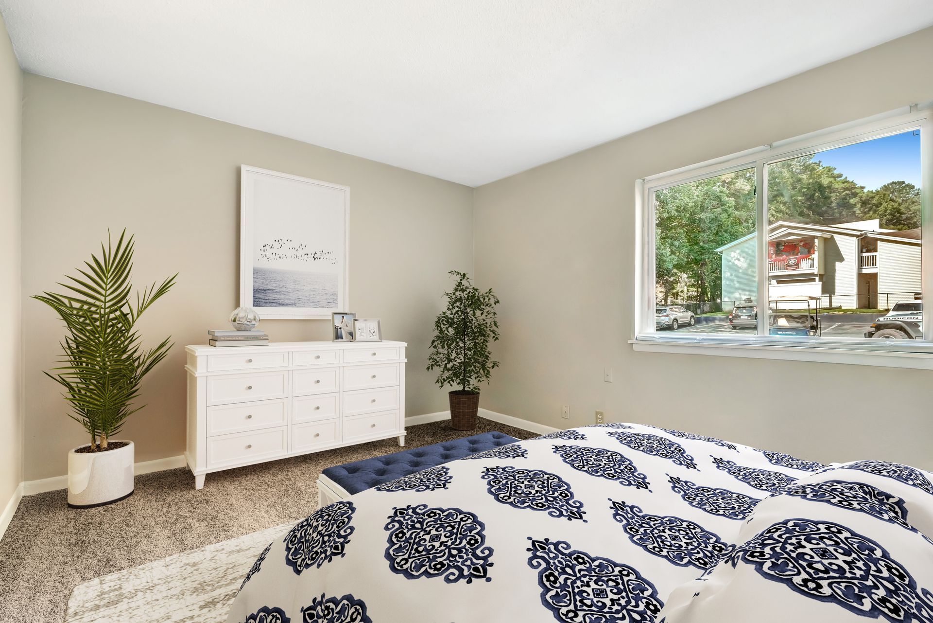 A furnished bedroom with a white dresser, two potted plants, and a bed with a blue and white patterned comforter.