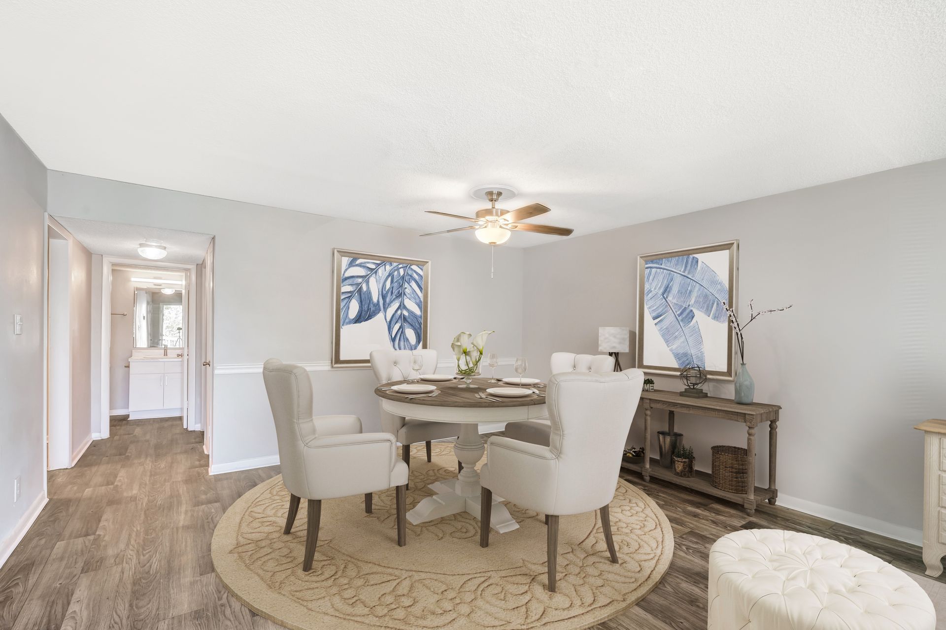 A dining area with a round table, four cream-colored chairs on a textured rug, two blue abstract art pieces, and a console.