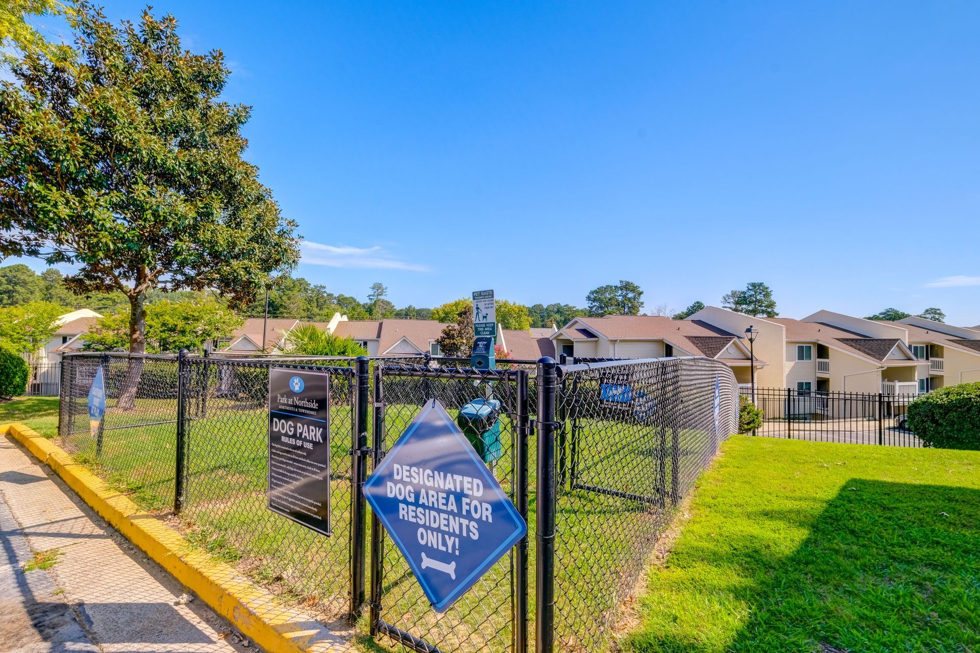 A fenced-in dog park with a blue sign stating the area is for residents only, set against an apartment complex.