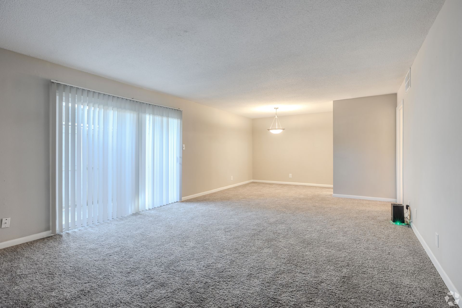 An empty living room with light gray carpet, beige walls, and a large sliding glass door with vertical blinds.