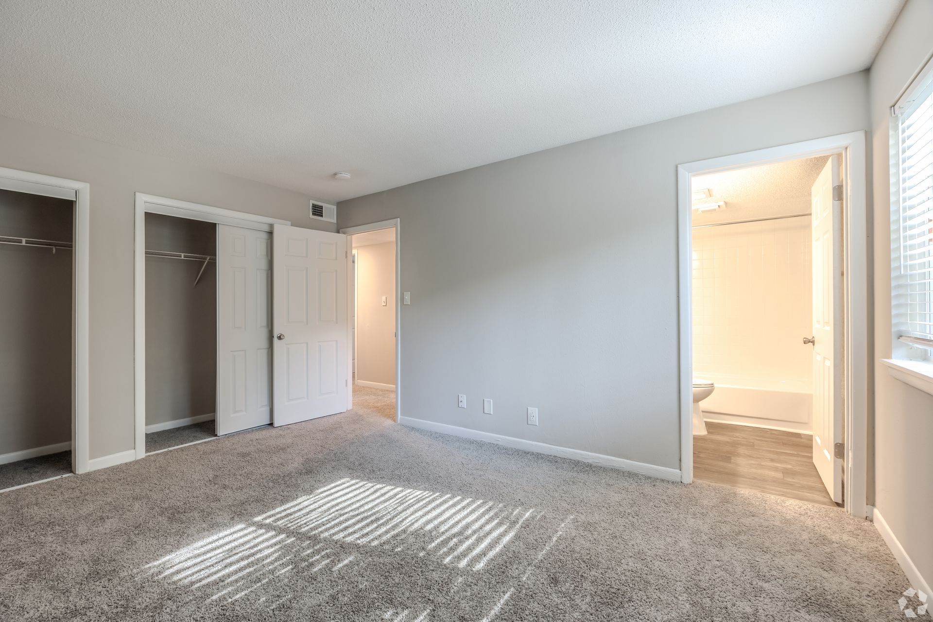A bedroom with gray carpet, light gray walls, two open closets, and an open doorway leading to a brightly lit bathroom.