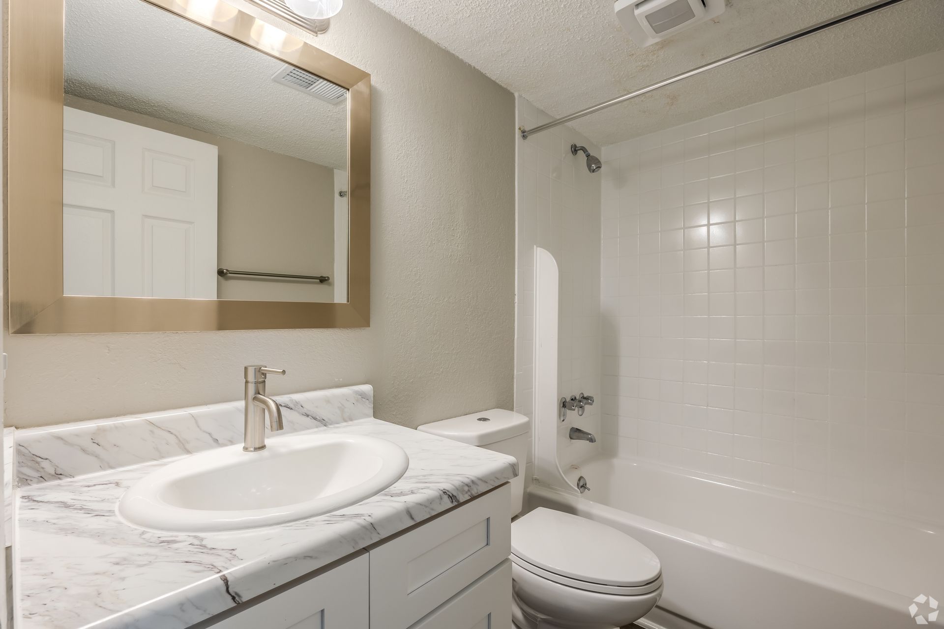 A bathroom with a white vanity, marble-patterned countertop, gold-framed mirror, toilet, and white shower-tub combination.