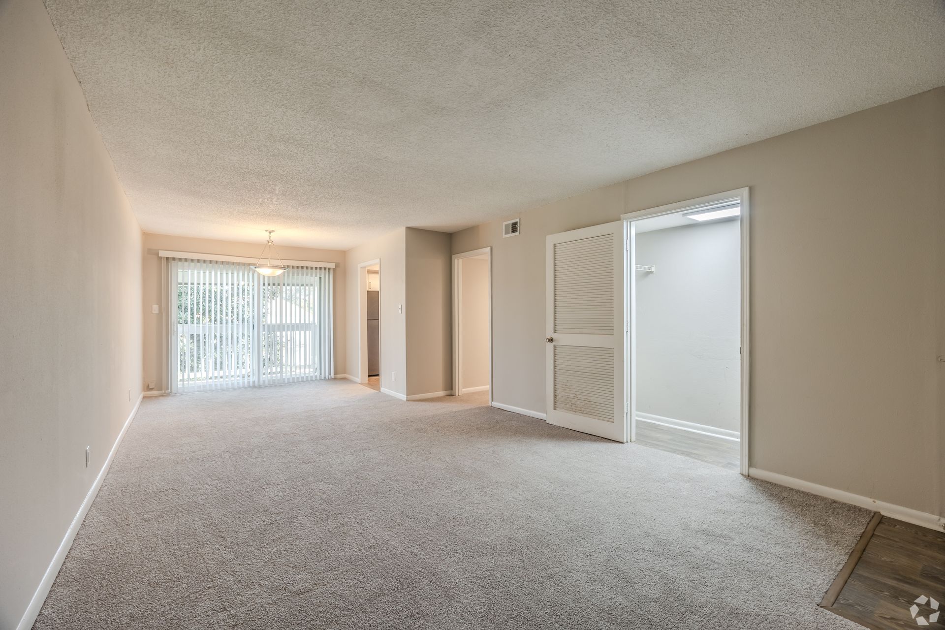 An empty apartment living room with beige carpet, light walls, a textured ceiling, and a sliding glass door to a balcony.
