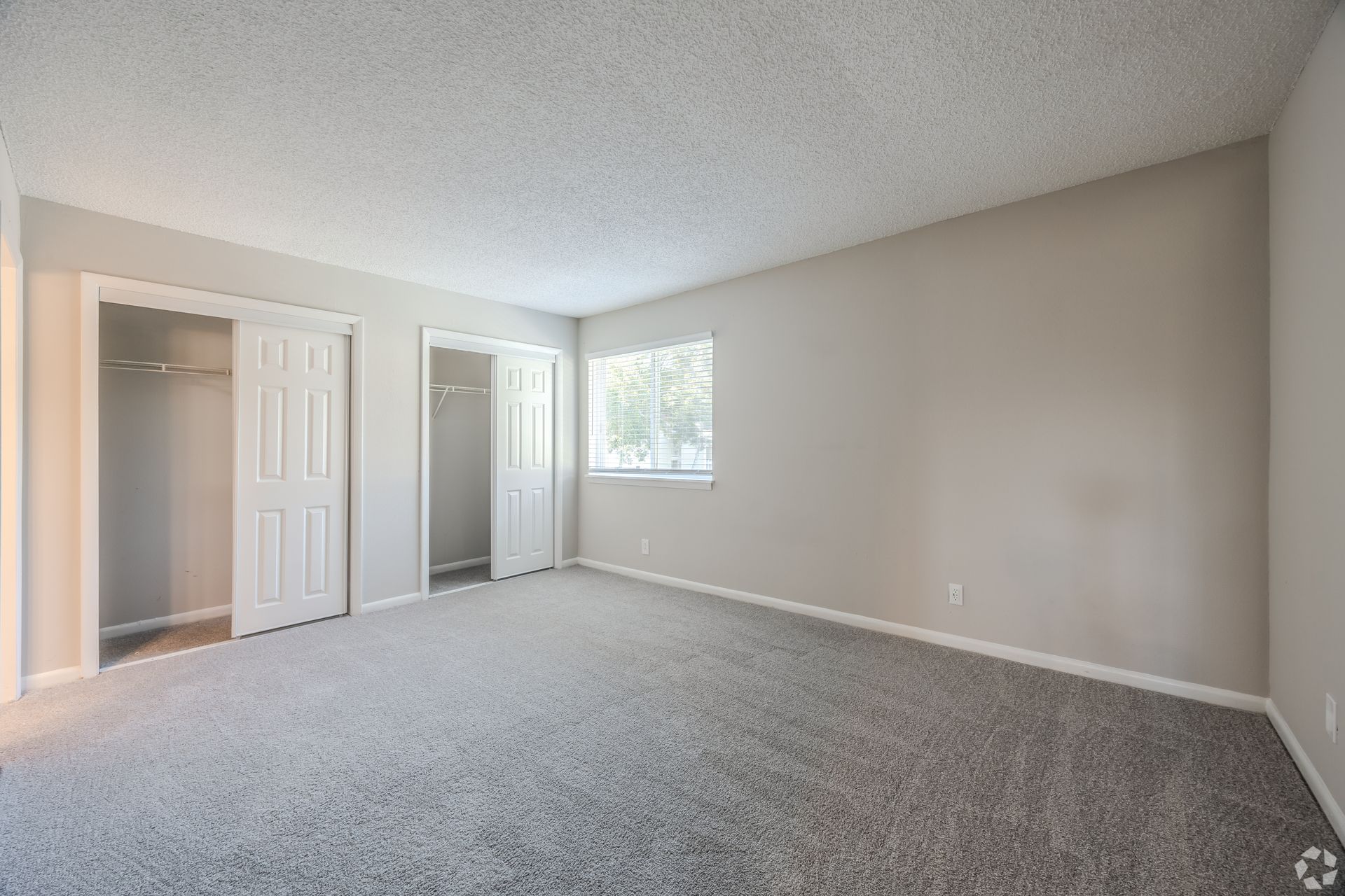 An empty bedroom with gray carpet, beige walls, a white ceiling with popcorn texture, and two white sliding-door closets.