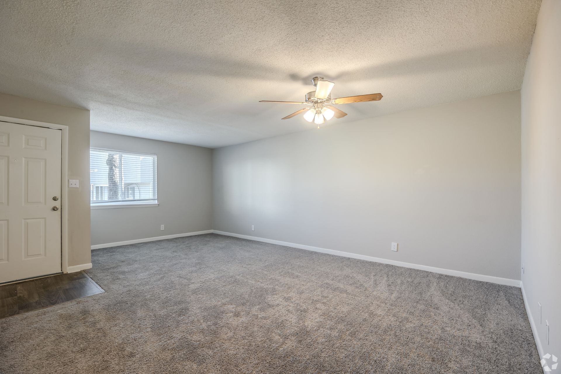 A spacious, empty living room with light gray walls, gray carpet, a ceiling fan, and a white entry door.