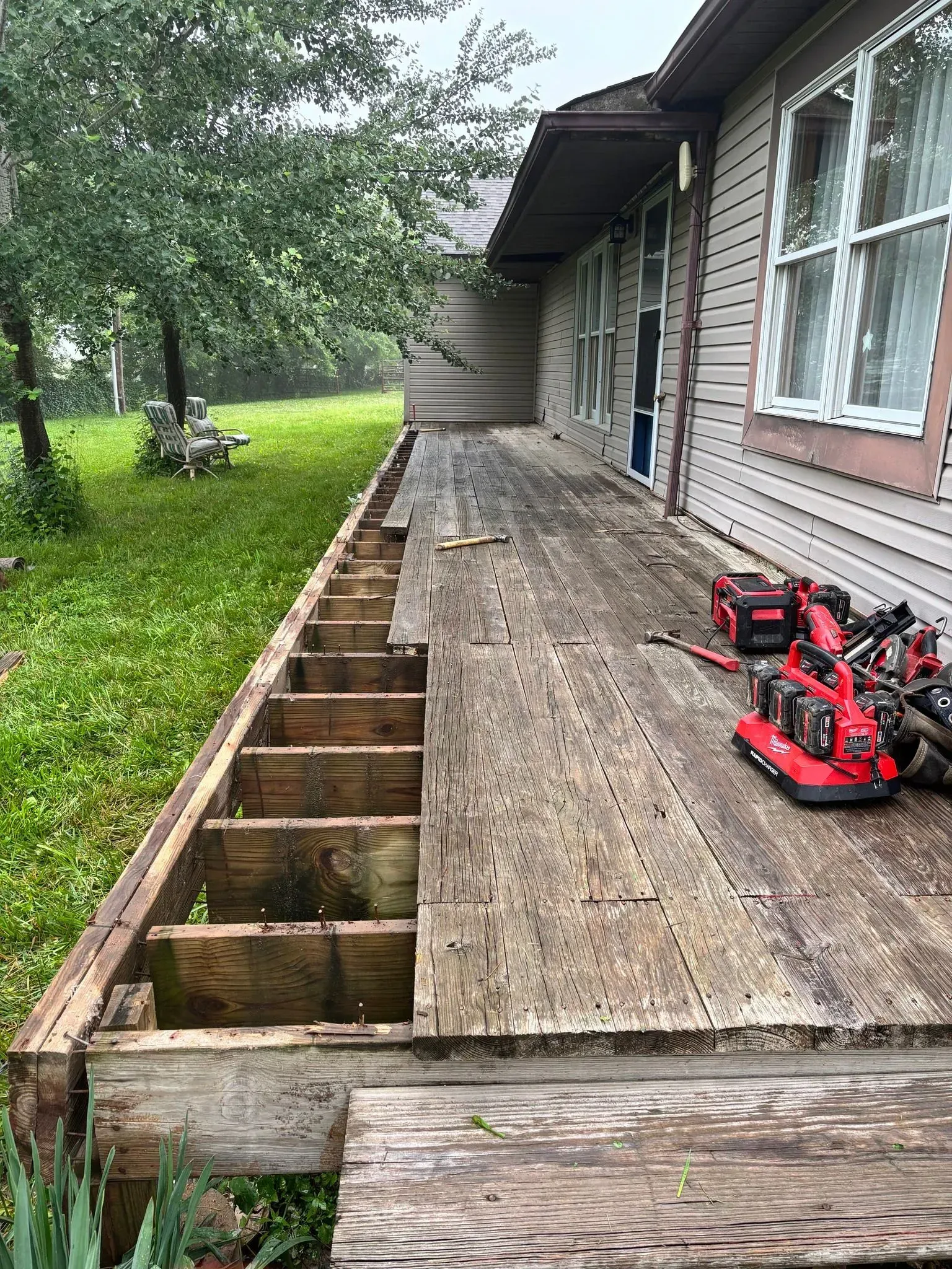 Partially dismantled wooden porch beside a house, with exposed joists and red tools on the deck.