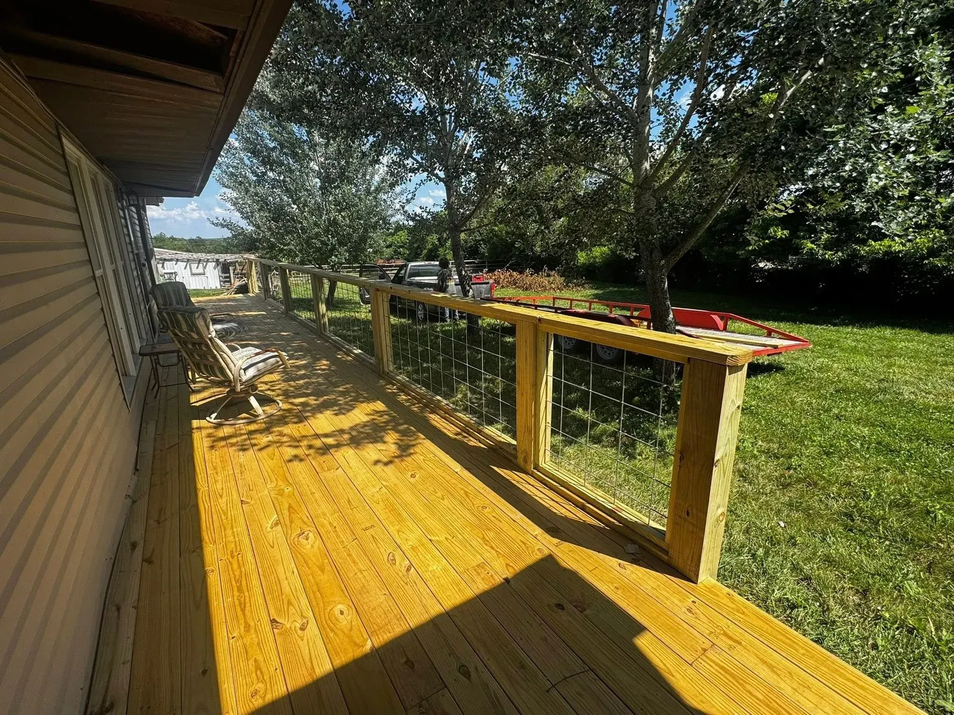 Wooden deck beside a house, shaded by trees, with yellow railing and red flowers along the edge.