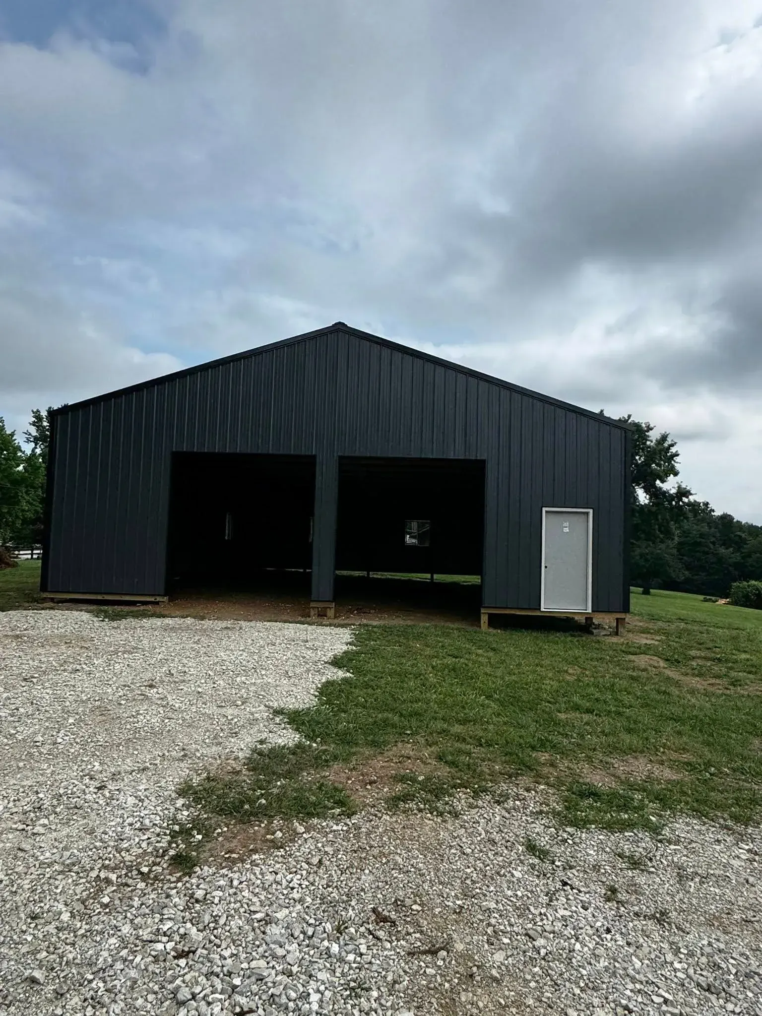Black metal barn with open bays and a white side door, beside a gravel drive under cloudy sky
