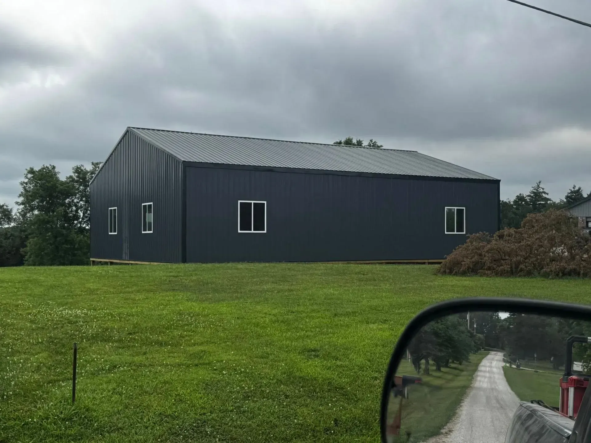 Dark blue barn-like building on a green field under cloudy skies, with a gravel road in the foreground