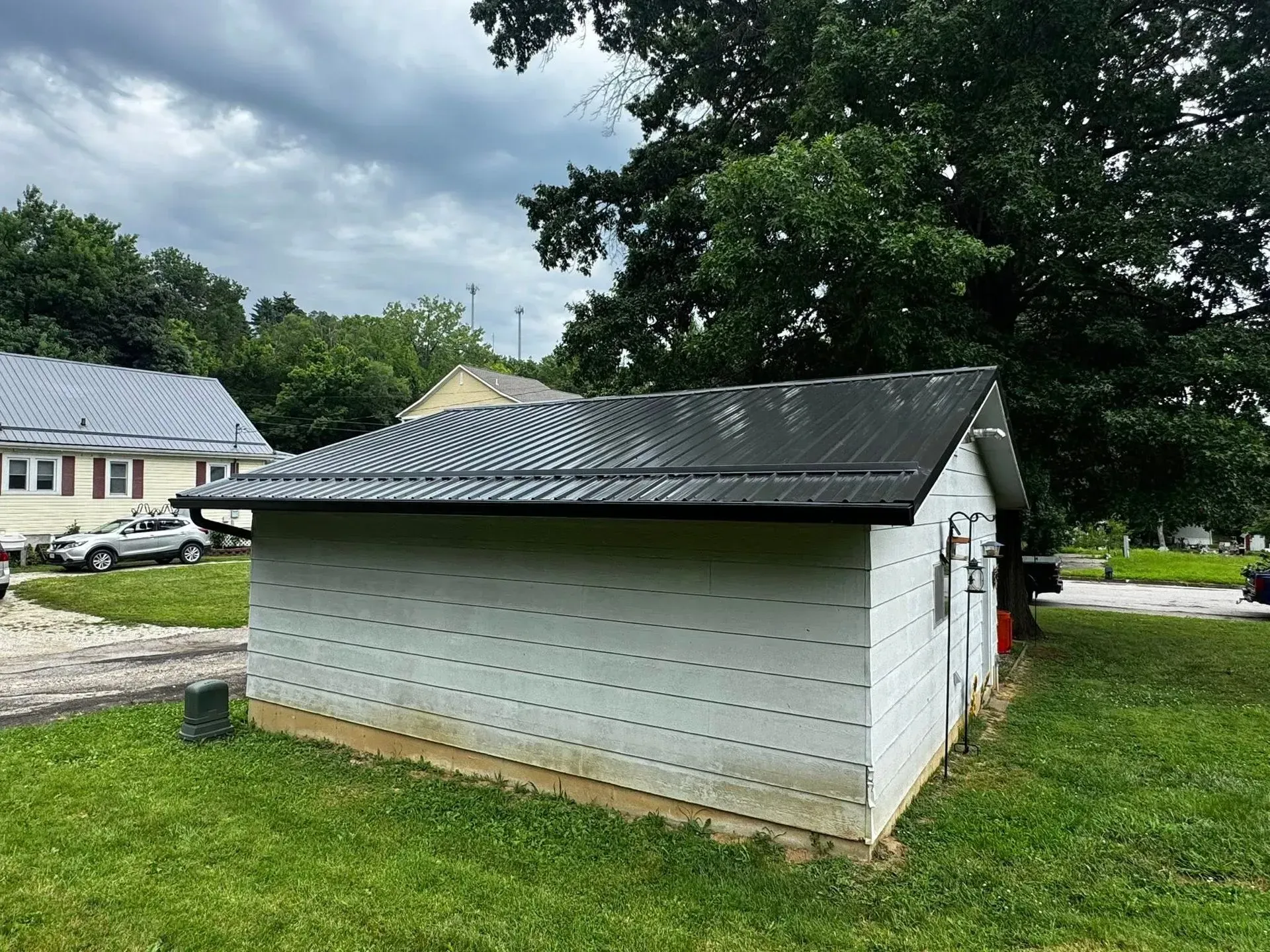 White utility shed with dark metal roof in a grassy yard, with houses and trees in the background.