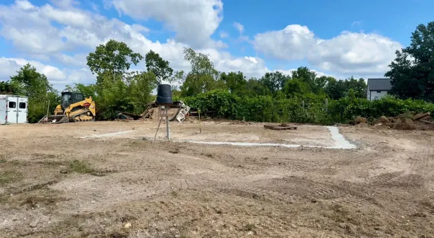 Construction site with excavator, dirt lot, and trees under a blue sky