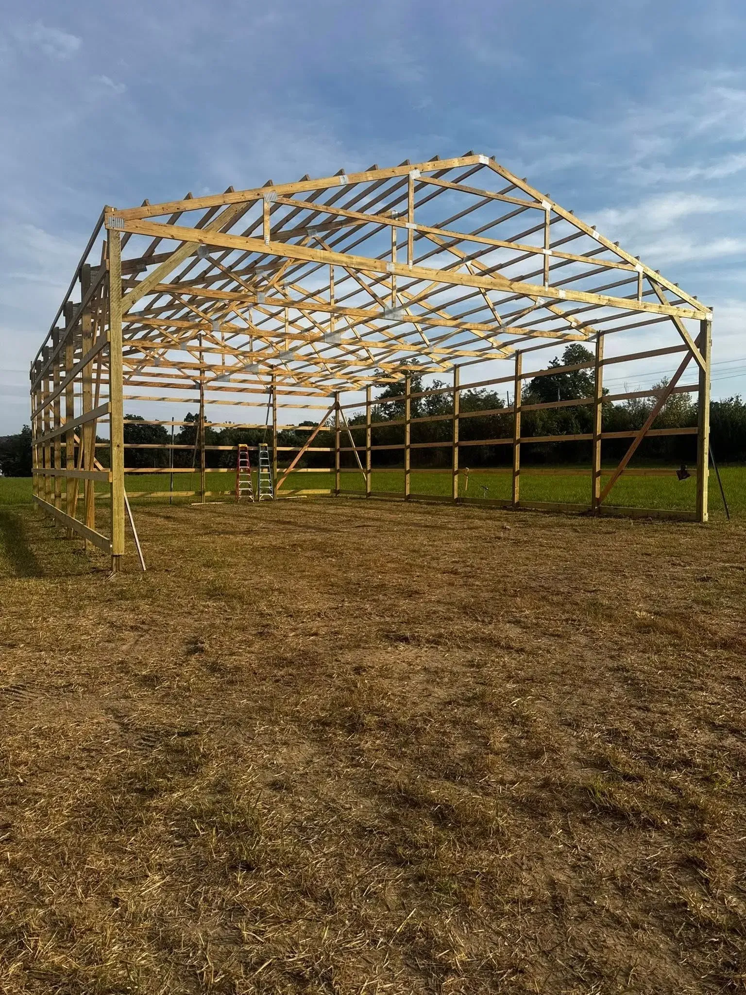 Partially framed wooden barn structure on a dirt field under a cloudy sky