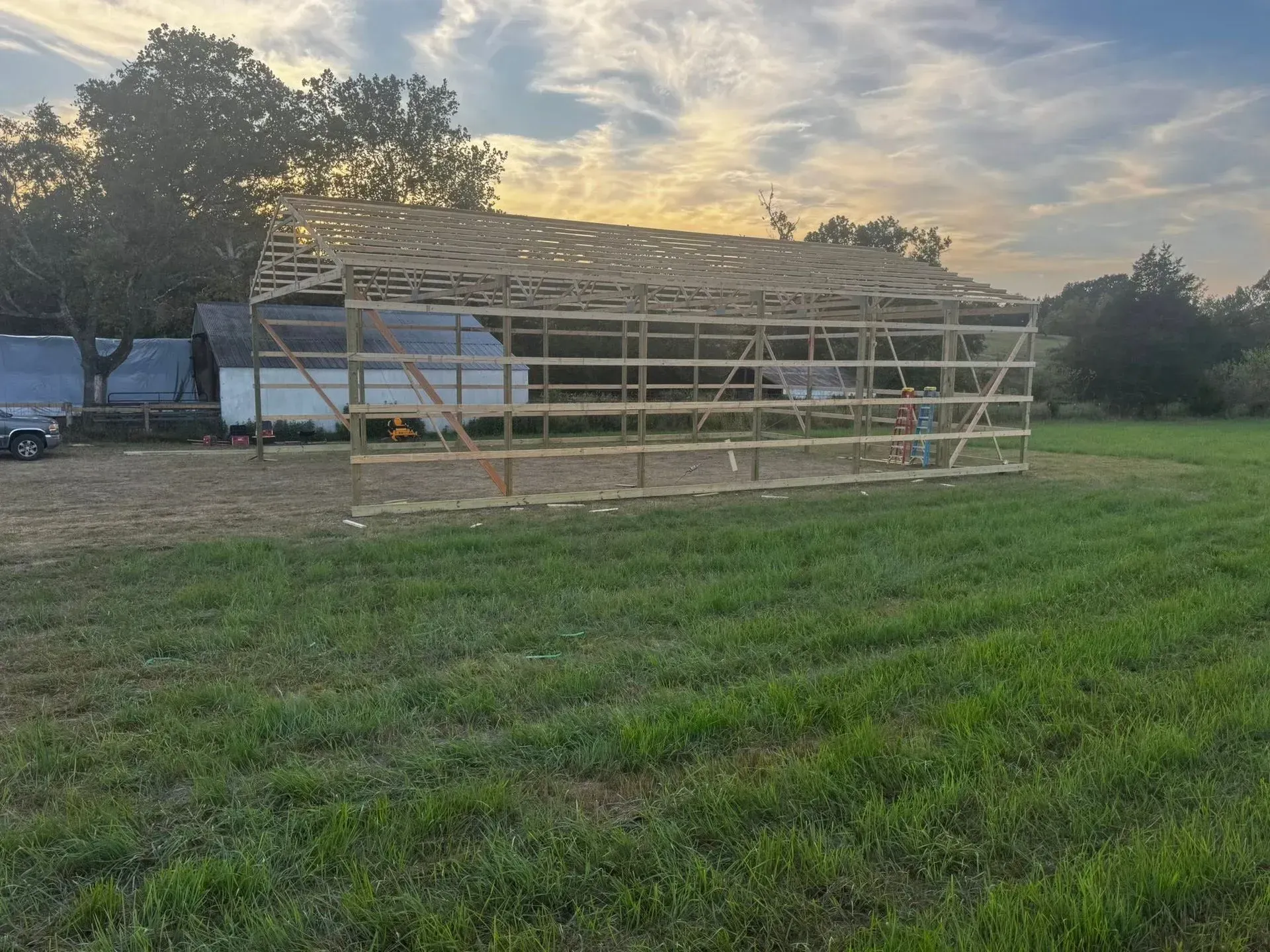 Metal barn frame in a grassy field at sunset, with trees and a trailer in the background.