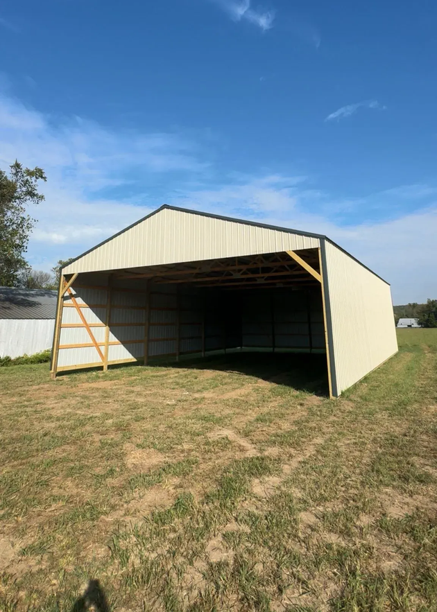 Open metal barn with tan siding and a wide front opening on a grassy field under a blue sky