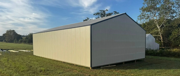 White utility shed with dark metal roof in a grassy yard, with houses and trees in the background.