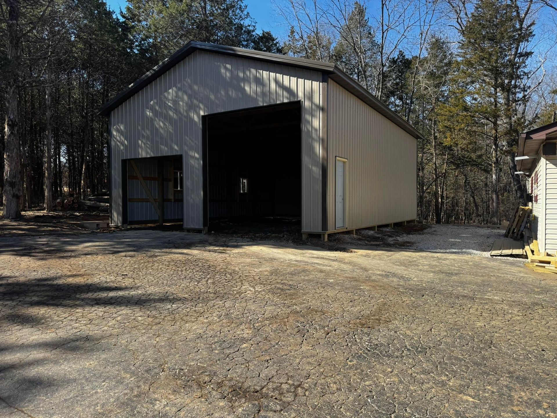 Beige metal garage with open bay doors on a gravel lot, surrounded by trees