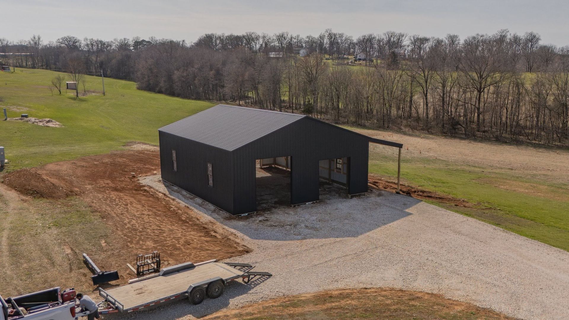 A black barn-style shed on a gravel drive beside a grassy field and trees.