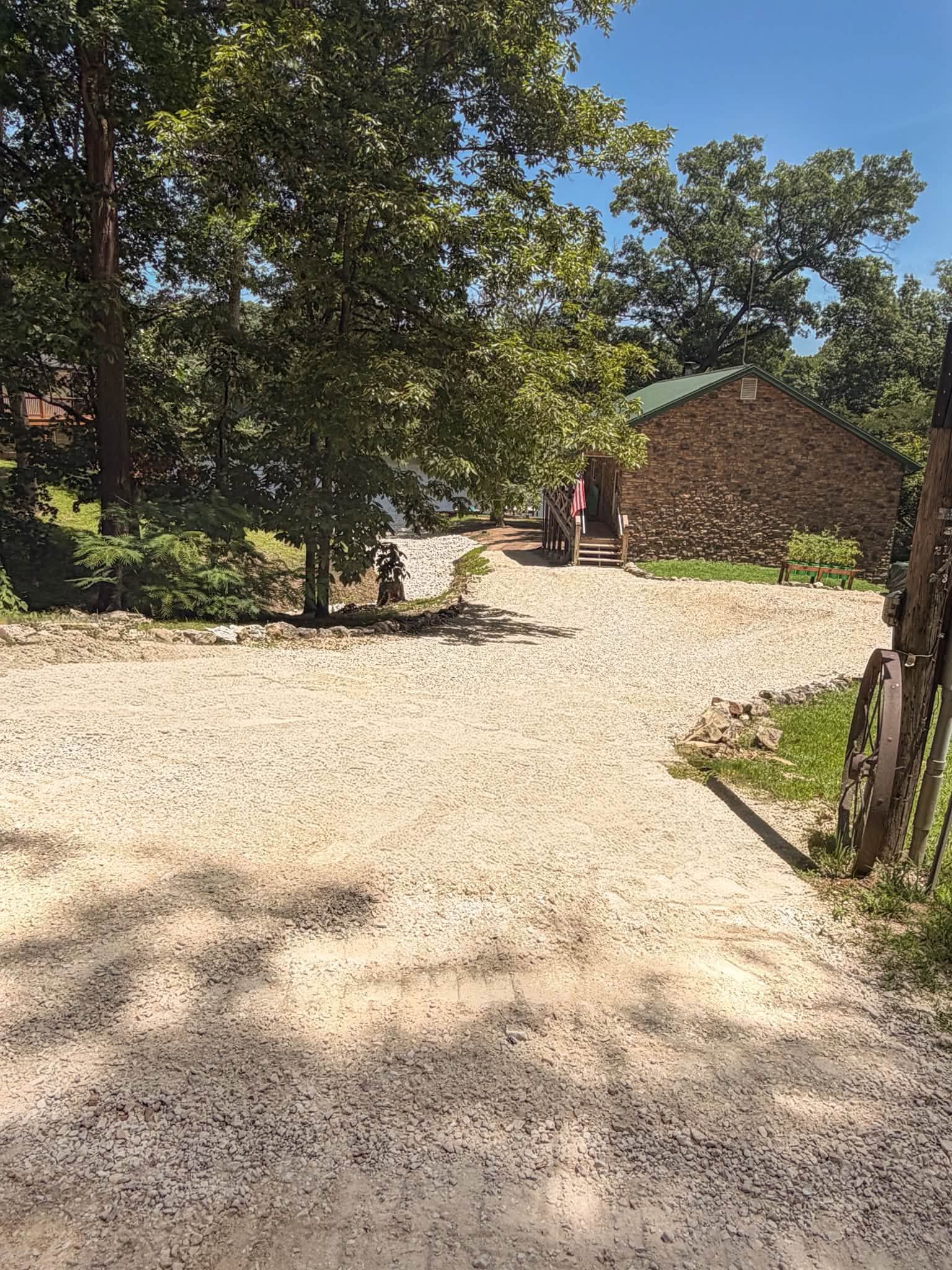 Gravel driveway beside trees, leading to a brick house under bright sunlight