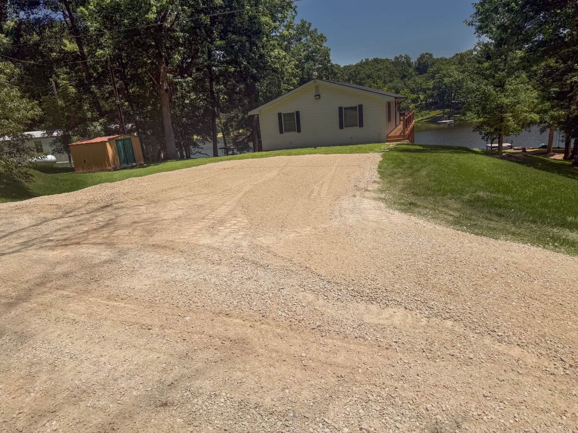 Gravel driveway leading to a small white house surrounded by trees and grass on a sunny day