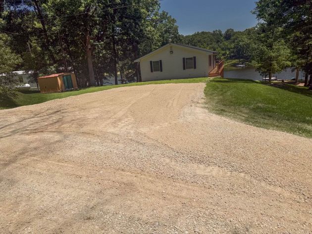 Gravel driveway leading to a small white house with trees and a lake in the background