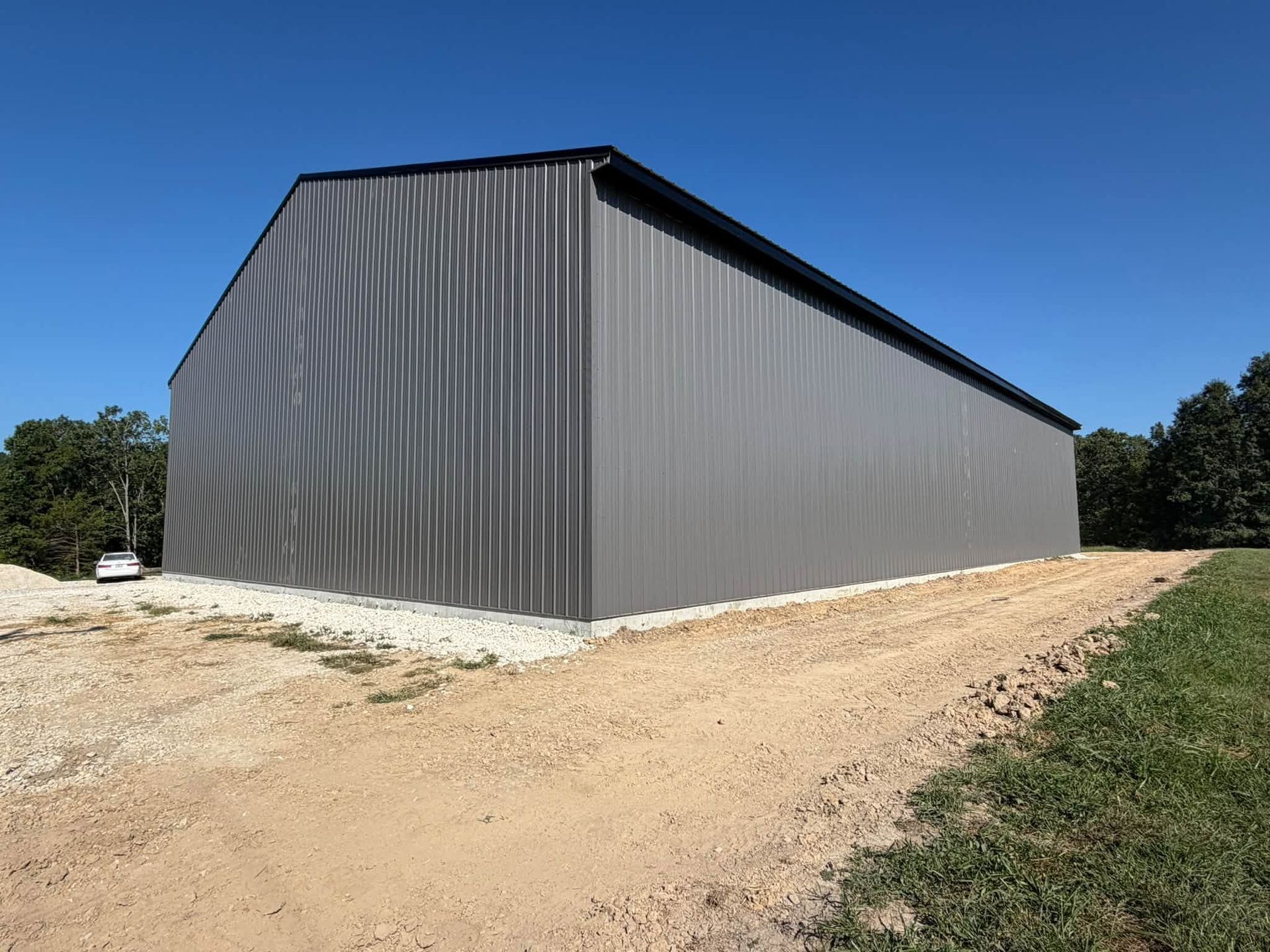 Large gray metal barn on a dirt road under a clear blue sky, with trees in the background