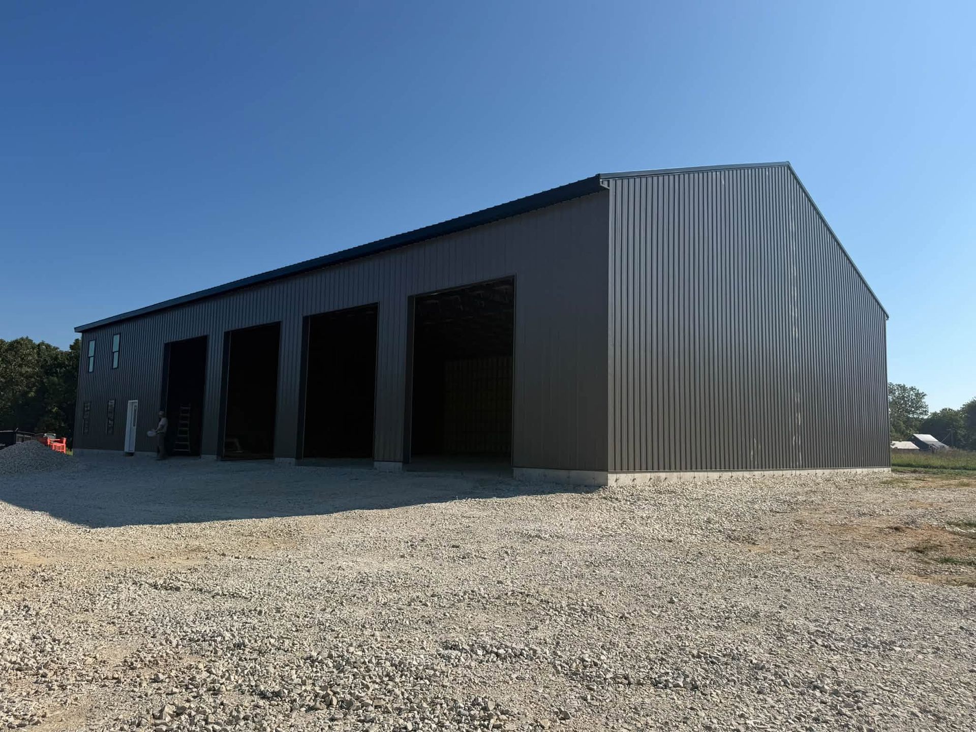 Large gray metal warehouse with open garage bays on a gravel lot under a clear blue sky