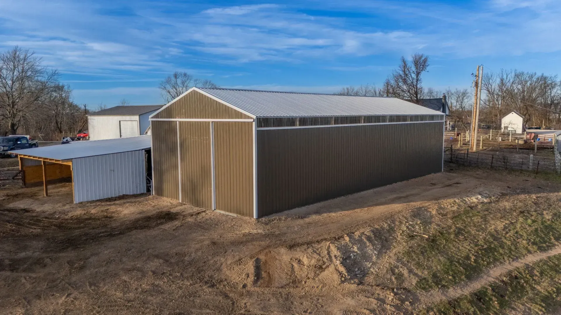 Large brown metal barn with a white roof in a rural dirt lot under a blue sky.