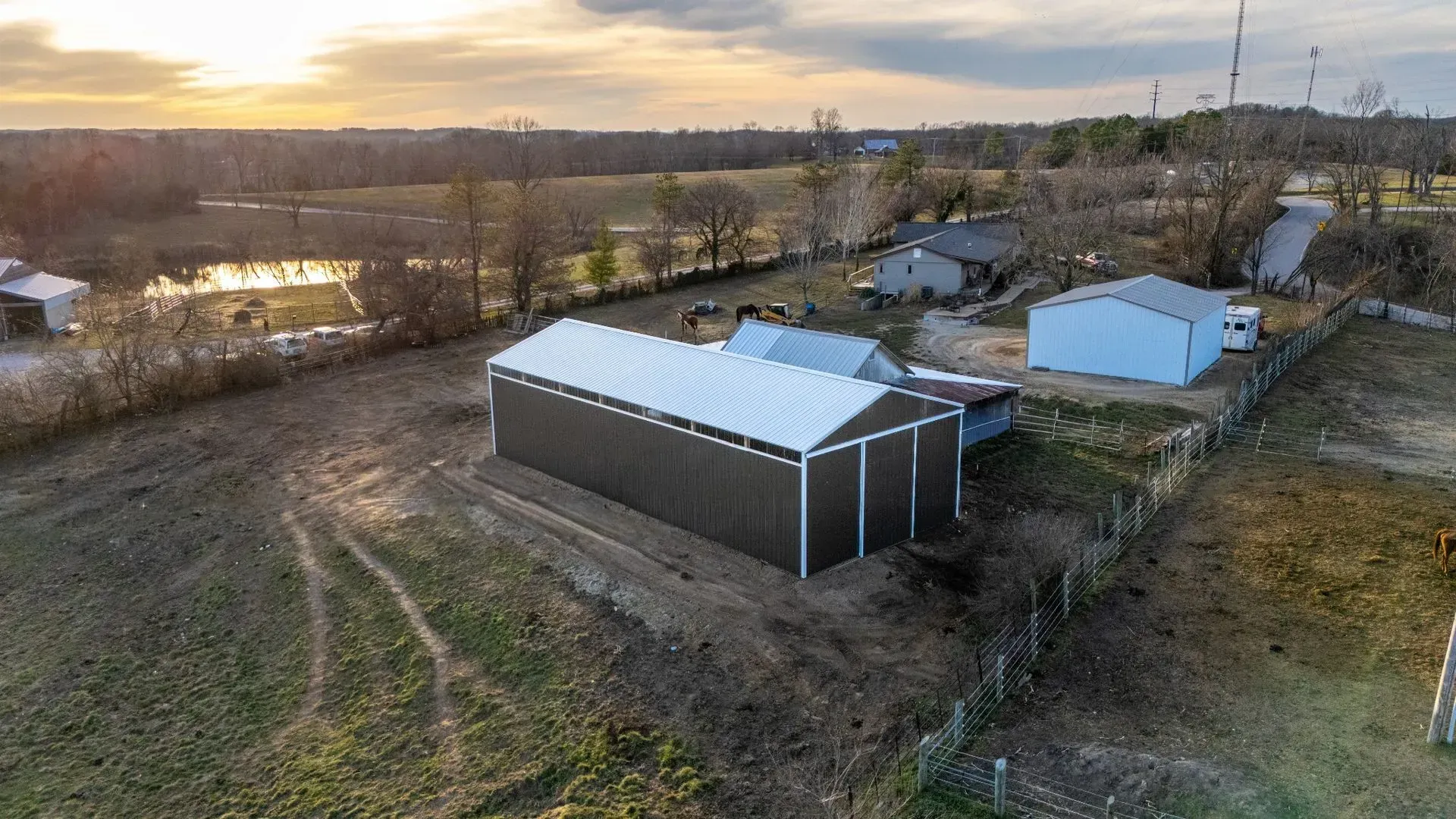 Rural farmyard at sunset with sheds, dirt tracks, and a river or flooded field in the background