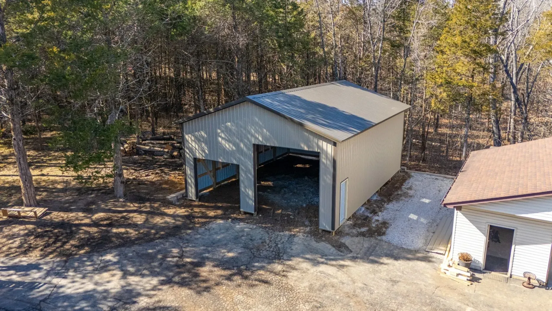 A white metal garage with open bays sits beside a small white shed in a wooded yard.