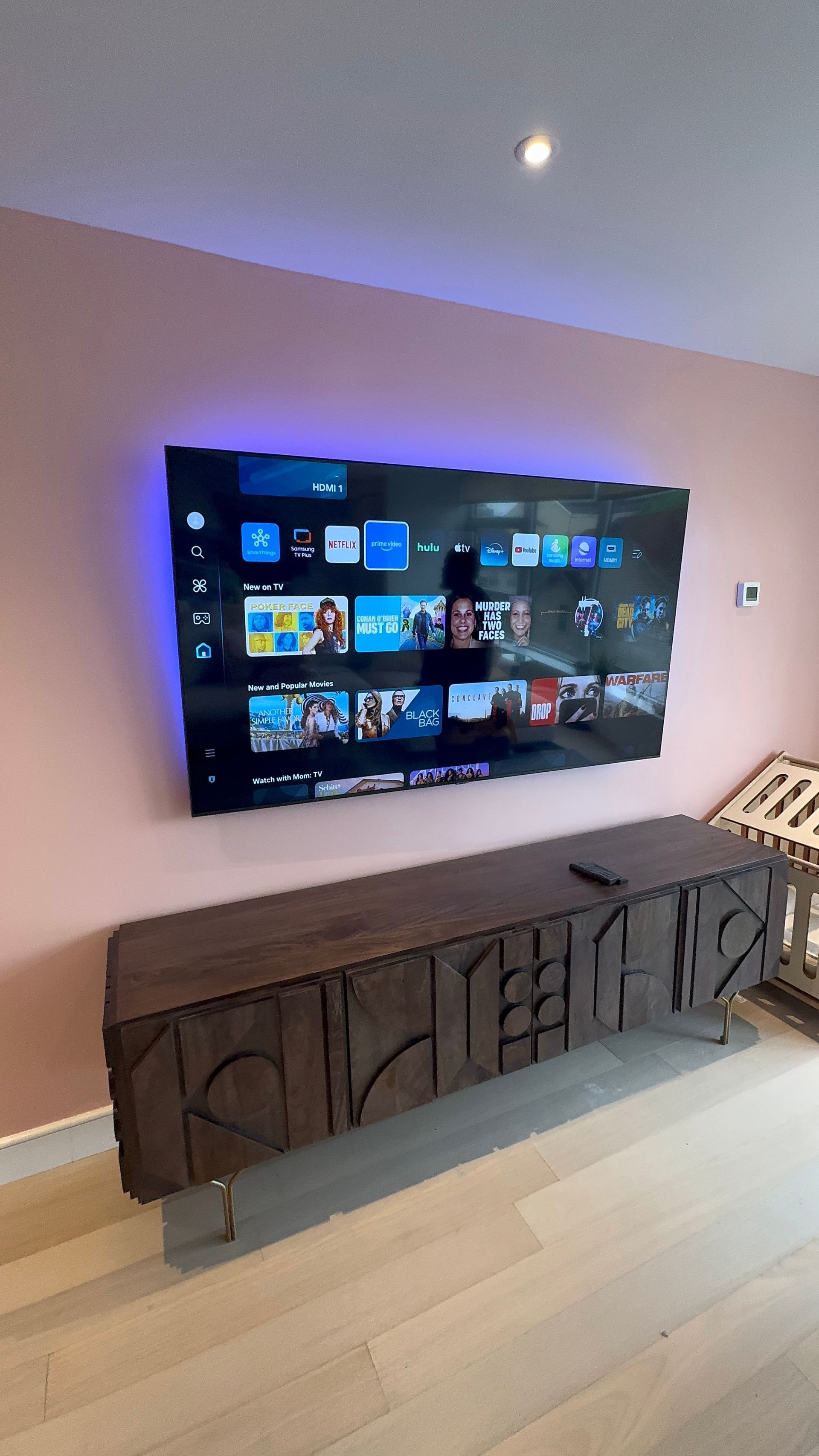 A wall-mounted TV with blue backlighting above a dark brown cabinet against a pink wall.