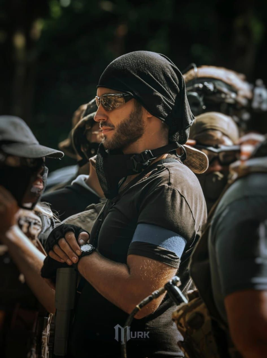 Man in black tactical gear and headscarf standing among others outdoors, holding his arms crossed