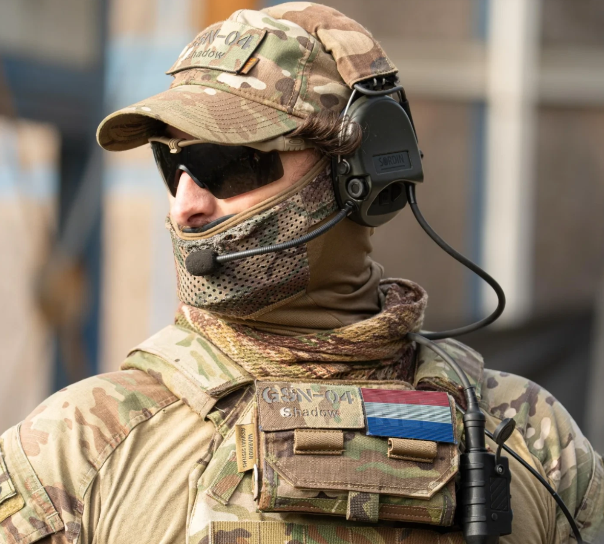 Camouflaged soldier wearing sunglasses, headset, and face covering, with a flag patch on tactical gear.