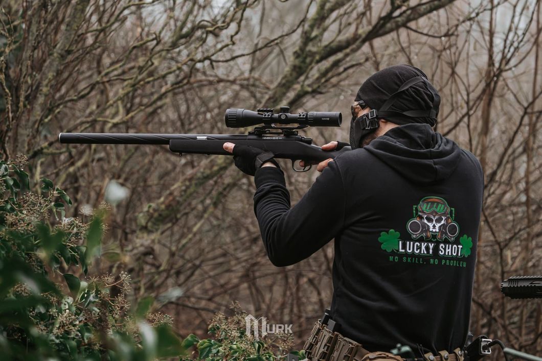 Person aiming a rifle with scope in a wooded outdoor setting, wearing a black hoodie.