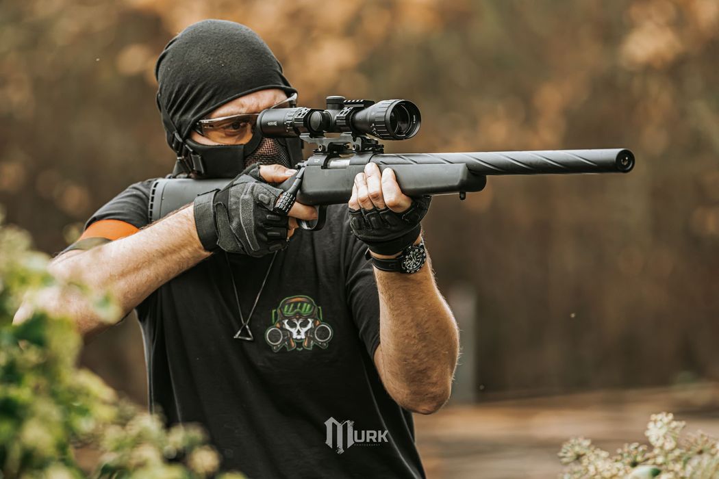 Masked person aiming a scoped rifle outdoors in a wooded area