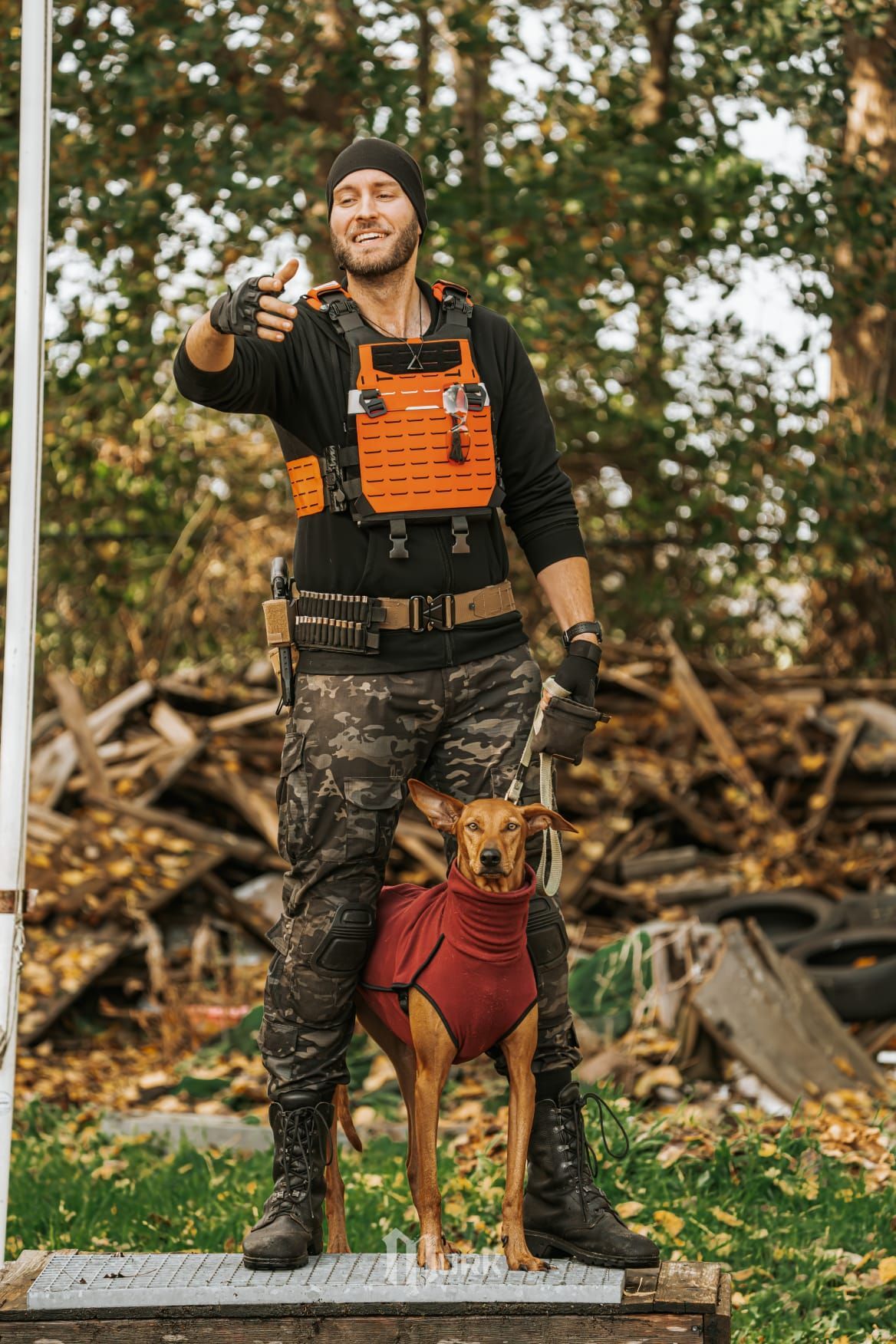Man in orange vest standing outdoors with a dog in a red harness, holding up a small object.