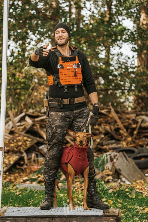 Man in orange vest standing outdoors with a dog in a red harness, holding up a small object.