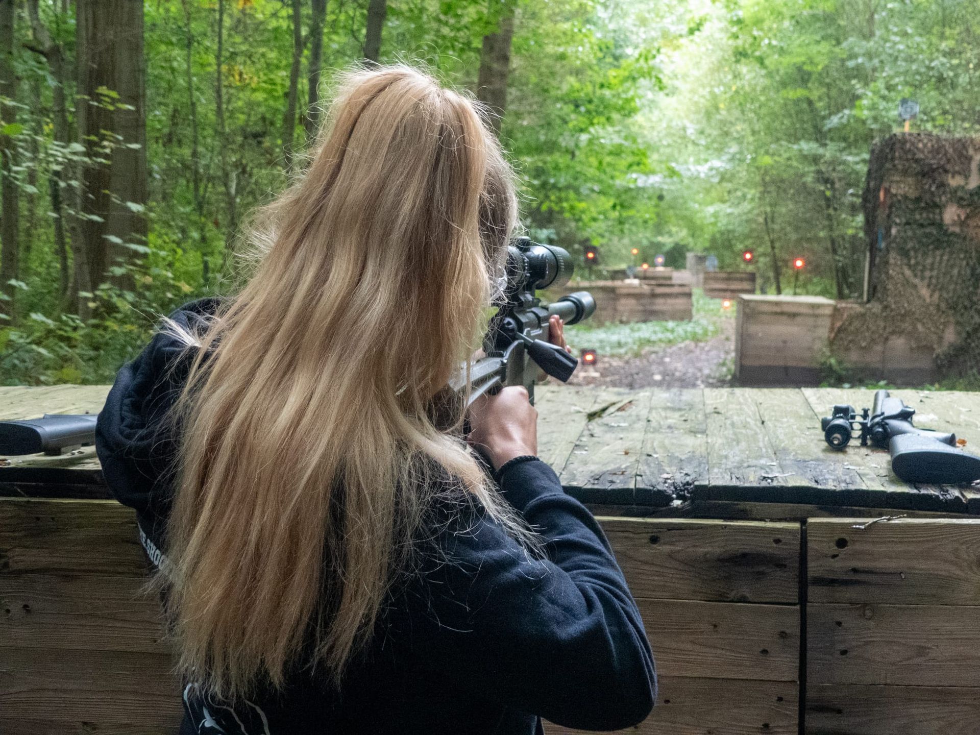Person aiming a rifle at an outdoor shooting range with targets ahead, surrounded by trees.