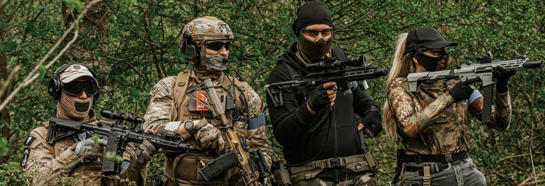 Four armed soldiers in camouflage advancing through a wooded area with rifles ready.