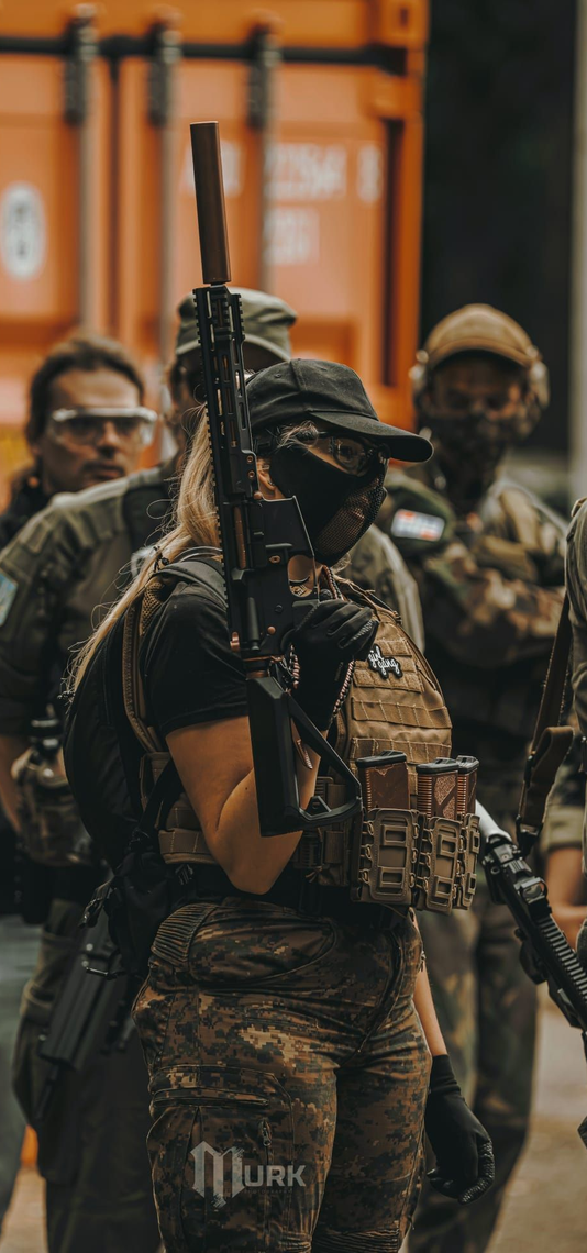 Armed tactical officers in camouflage gear standing indoors, one holding a rifle upright.