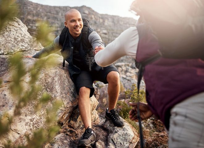 Hiker smiling while climbing a rocky mountain trail and reaching for a helper’s hand Hiker smiling while climbing a rocky mountain trail and reaching for a helper’s hand