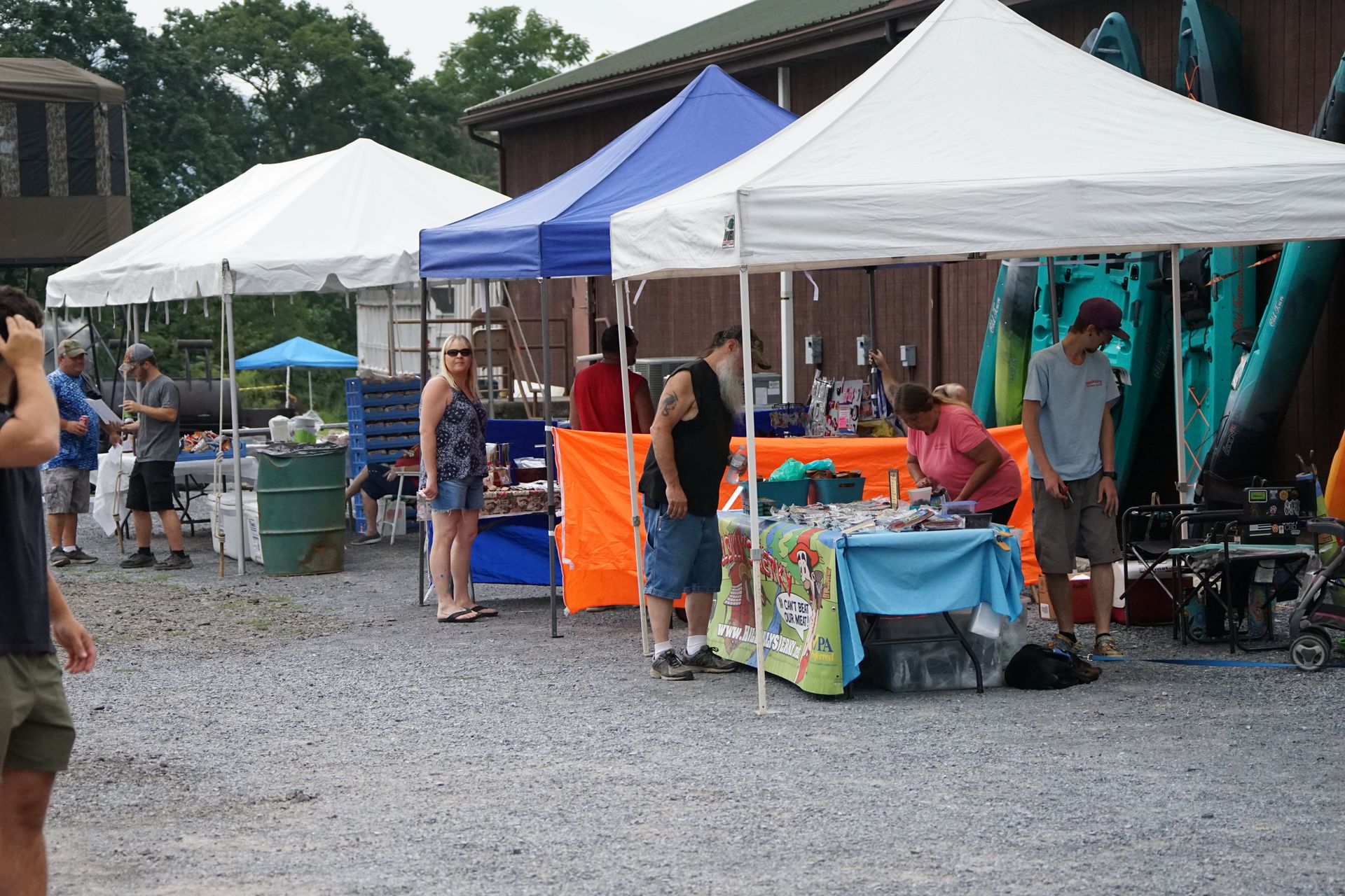 Outdoor market stalls under white and blue tents with people browsing on a gravel lot.