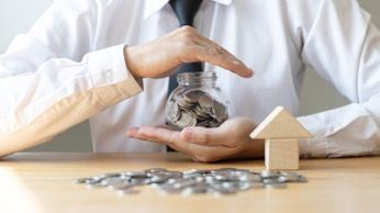 Hands covering a glass jar of coins beside a small house model on a table