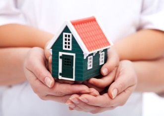 Hands gently holding a small green toy house with a red roof