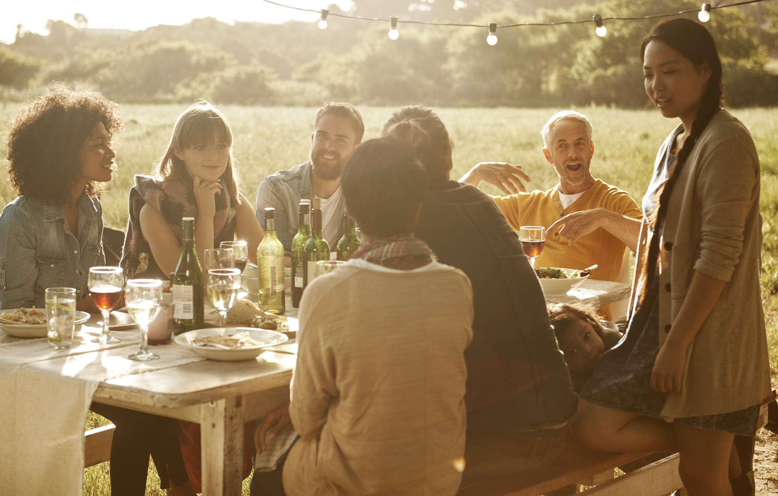 People at outdoor table, dining. Food, drinks, and string lights on a sunny day.