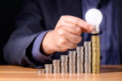 Person holding a lightbulb over a stacked coin graph, representing financial growth and ideas.