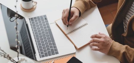 Person writes in a notebook with a laptop and coffee cup on a white desk.