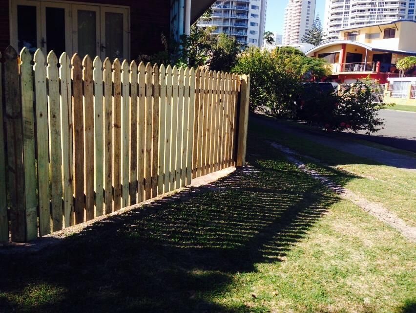 Wooden picket fence along a grassy lawn beside a house in bright sunlight.